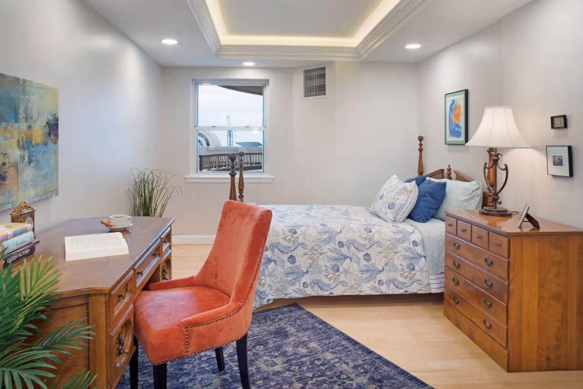 Well-lit bedroom with a wooden bed and dresser, a patterned blue bedspread, a wooden desk with a red upholstered chair, and a window.