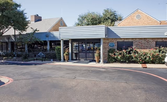 Exterior view of a single-story brick building with a covered entrance, potted plants on either side of the door, and landscaping with bushes and trees. The sky is clear and the pavement is visible in front of the building.