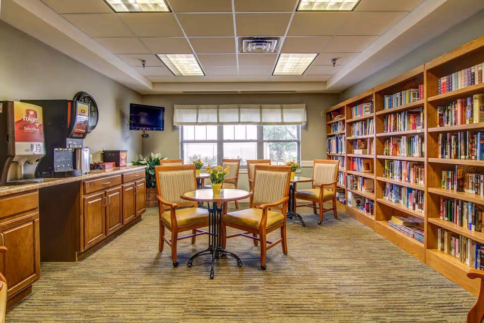 A cozy room with wooden bookshelves filled with books on the right side, several small round tables with chairs arranged around them, and a coffee and beverage station on the left side. The room has a large window with white curtains letting in natural light, a wall-mounted TV, and a carpeted floor.