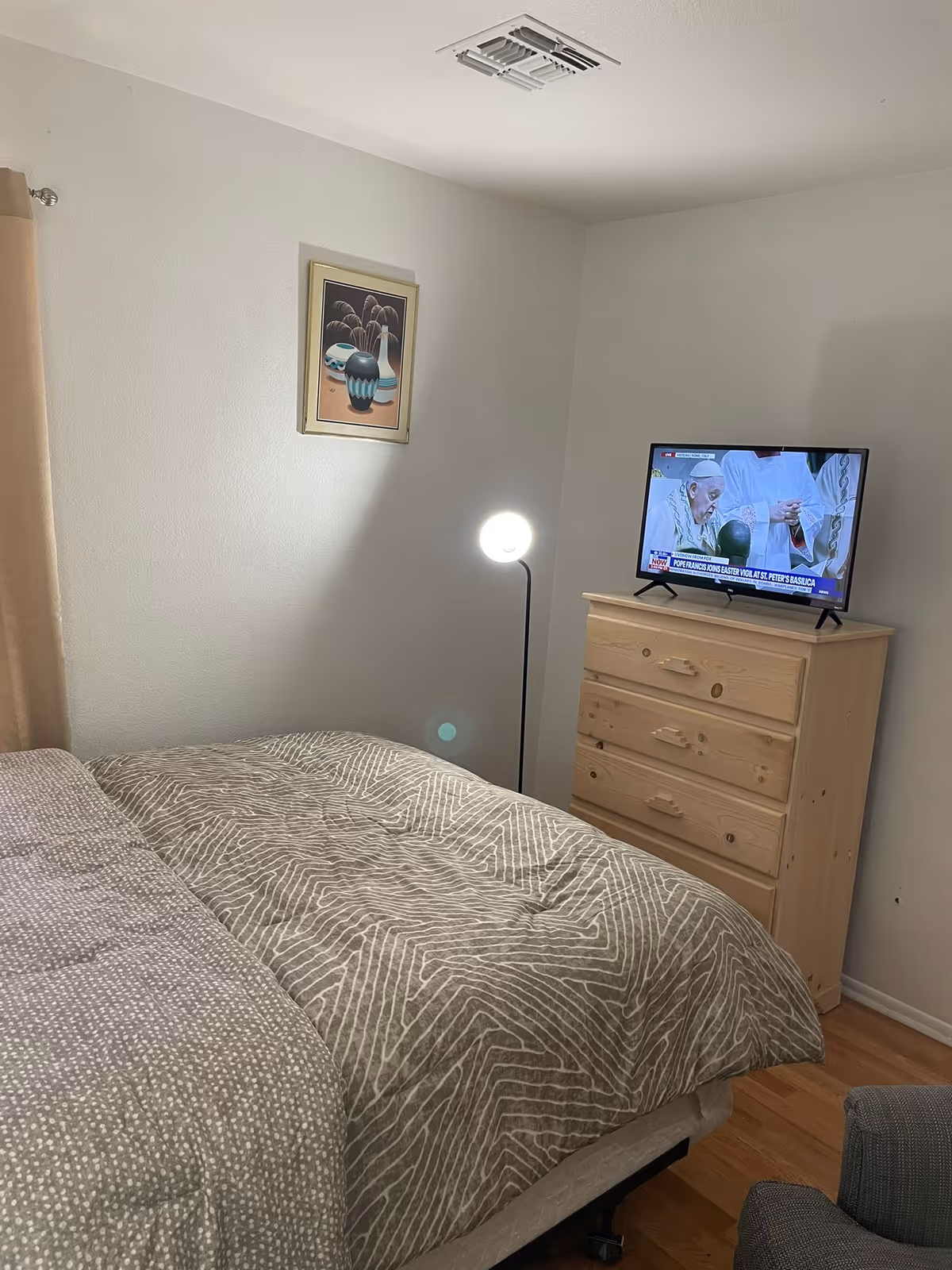 A bedroom with a bed covered in a patterned gray and white comforter, a wooden dresser with a TV on top showing a religious broadcast, a standing lamp emitting light, a framed picture on the wall, and beige curtains partially visible on the left side.