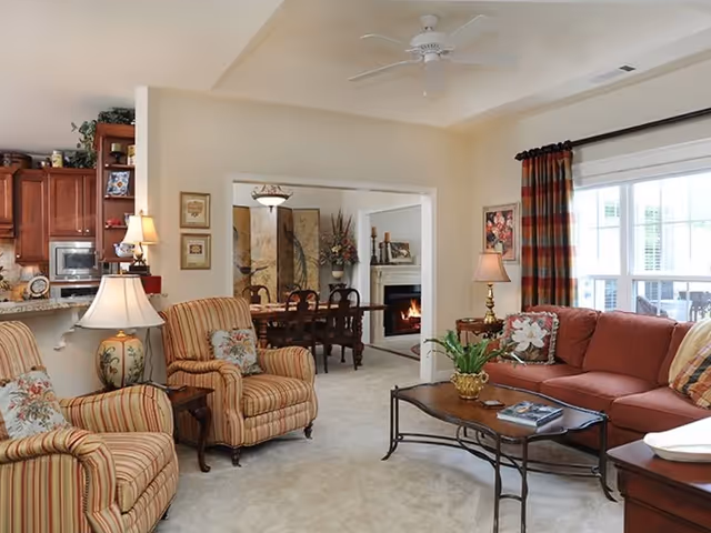A cozy living room with a red sofa, two striped armchairs, a wooden coffee table with a plant and books, and two table lamps. The room has a large window with striped curtains letting in natural light. In the background, there is a dining area with a wooden dining table and chairs, a fireplace with a fire burning, and decorative artwork on the walls.