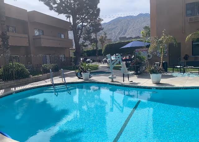 Outdoor swimming pool area with clear blue water surrounded by a concrete deck. There are potted plants, a pool lift, and patio tables with umbrellas. Residential buildings and mountains are visible in the background under a partly cloudy sky.