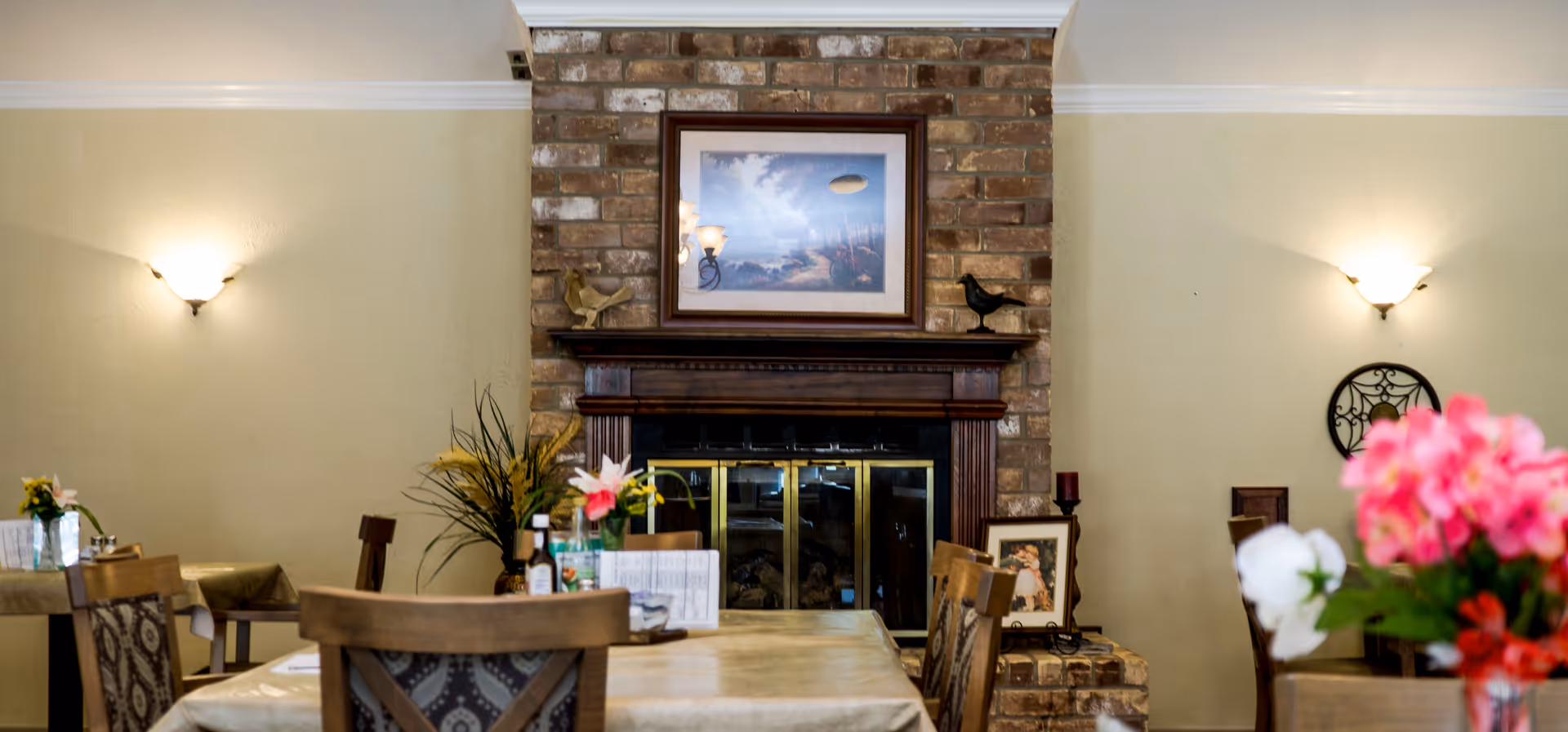 Interior view of a dining area with tables and chairs arranged around a fireplace made of brick. The fireplace has a wooden mantle with decorative items and a framed picture above it. The walls are painted beige with two wall-mounted light fixtures on either side of the fireplace. There are flowers placed on the tables in the foreground.
