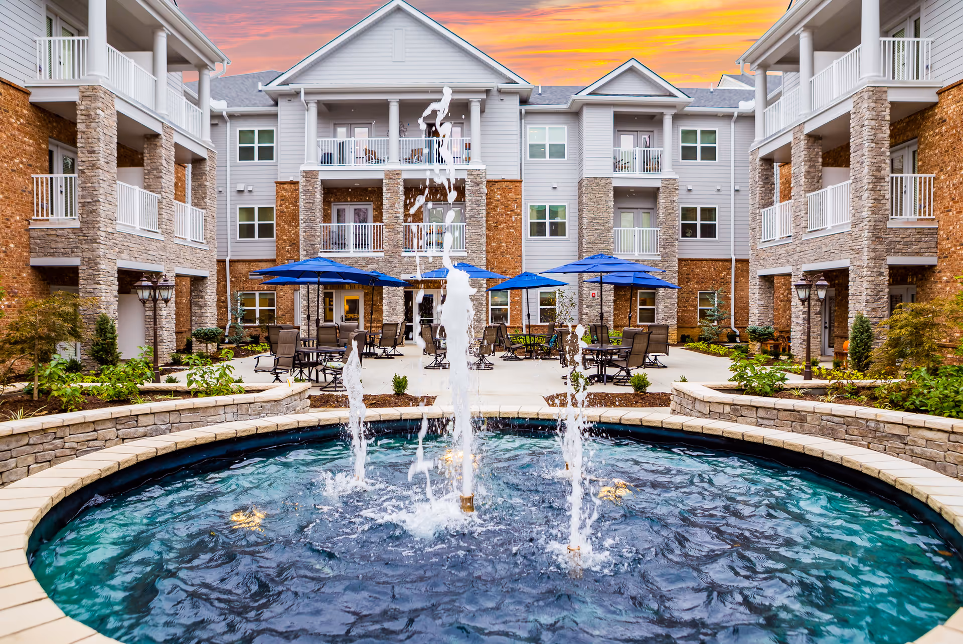 Courtyard of a senior living facility featuring a central fountain, patio tables with blue umbrellas, and surrounding multi-story residential buildings.