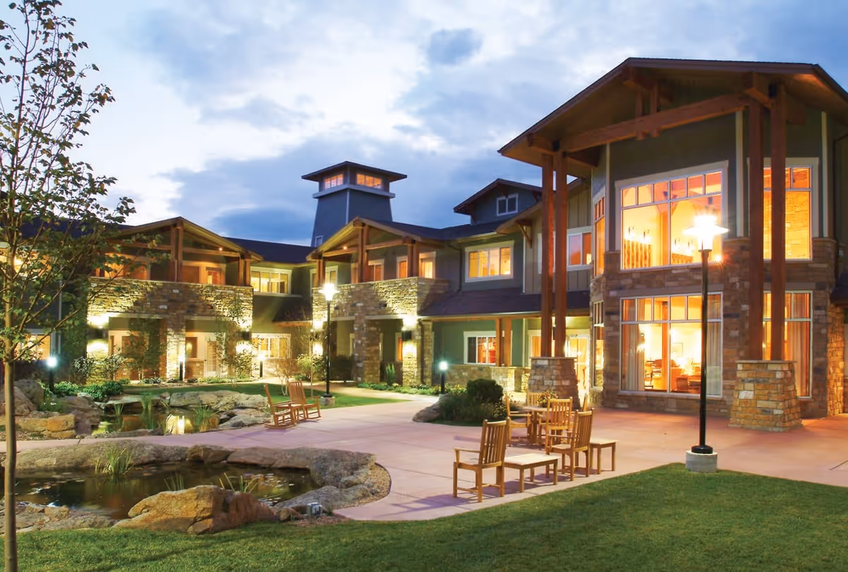 Exterior view of Balfour Louisville Campus at dusk, showing a well-lit building with large windows, stone and wood architectural details, outdoor seating with wooden chairs and tables, a small pond with rocks, and landscaped greenery.