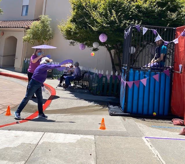 An outdoor scene at Cadence Millbrae showing a person throwing a ball towards a dunk tank where another person is seated inside. Two other people are sitting on a bench in the background near a picket fence and trees. The area is decorated with purple and pink paper lanterns and bunting.