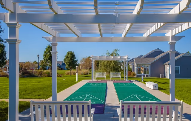 Outdoor shuffleboard courts with green playing surfaces under white pergolas, surrounded by green grass and trees, with benches nearby and residential buildings in the background under a clear blue sky.