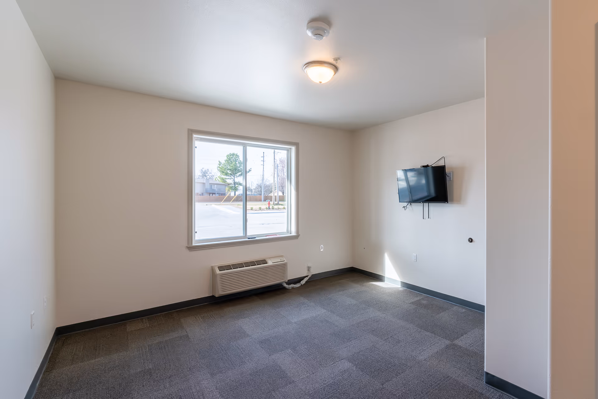 Empty room with gray carpet, white walls, a window with a view outside, a wall-mounted TV, and a ceiling light fixture.