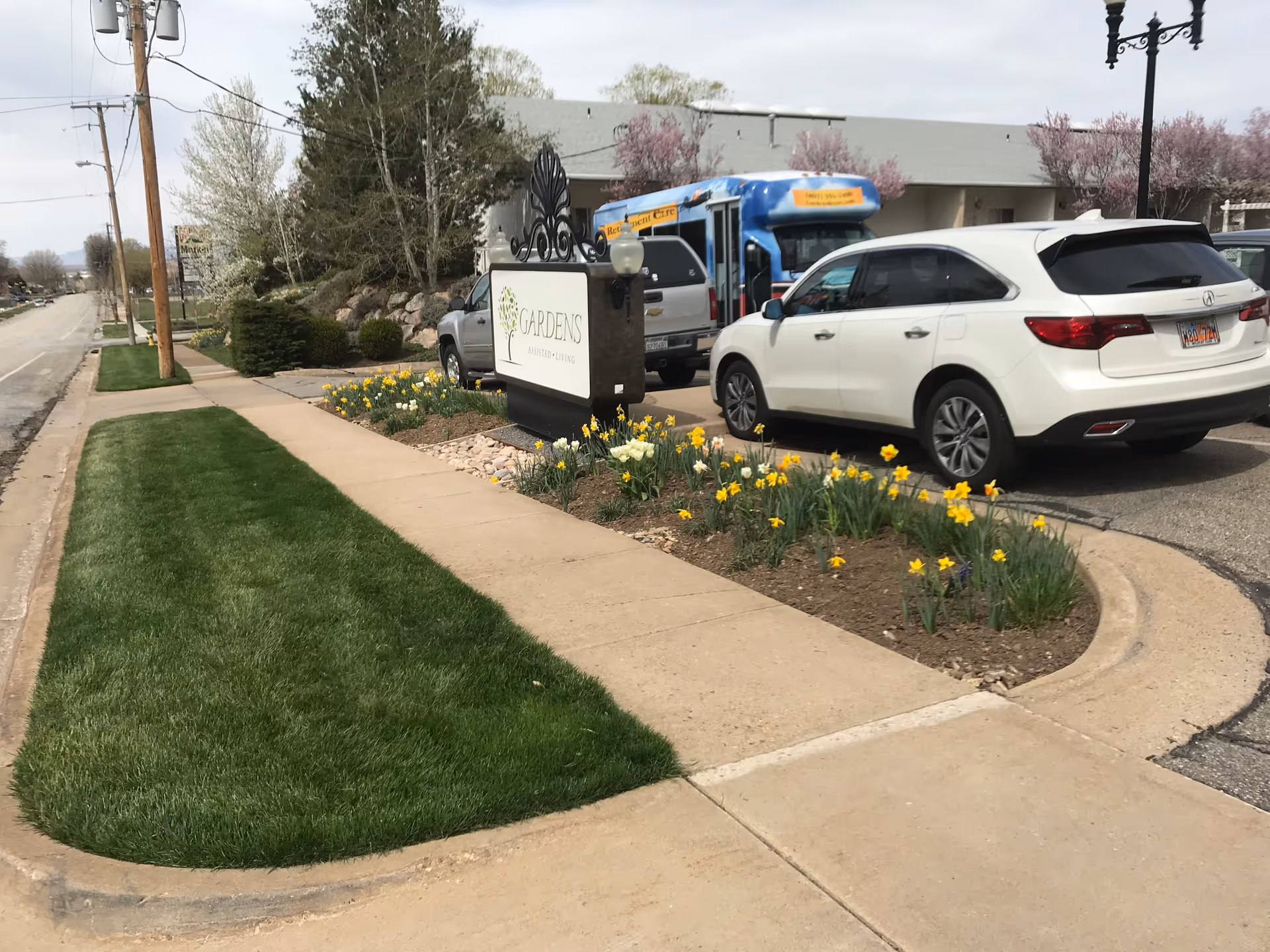 Sidewalk and landscaped area with yellow flowers in front of a sign for Gardens Assisted Living, with parked cars and a blue bus in the background.