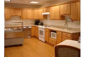 A spacious kitchen with wooden cabinets, white appliances including a stove, microwave, and dishwasher, and a countertop with a sink. The floor is made of light wood, and there is a chair partially visible in the foreground.