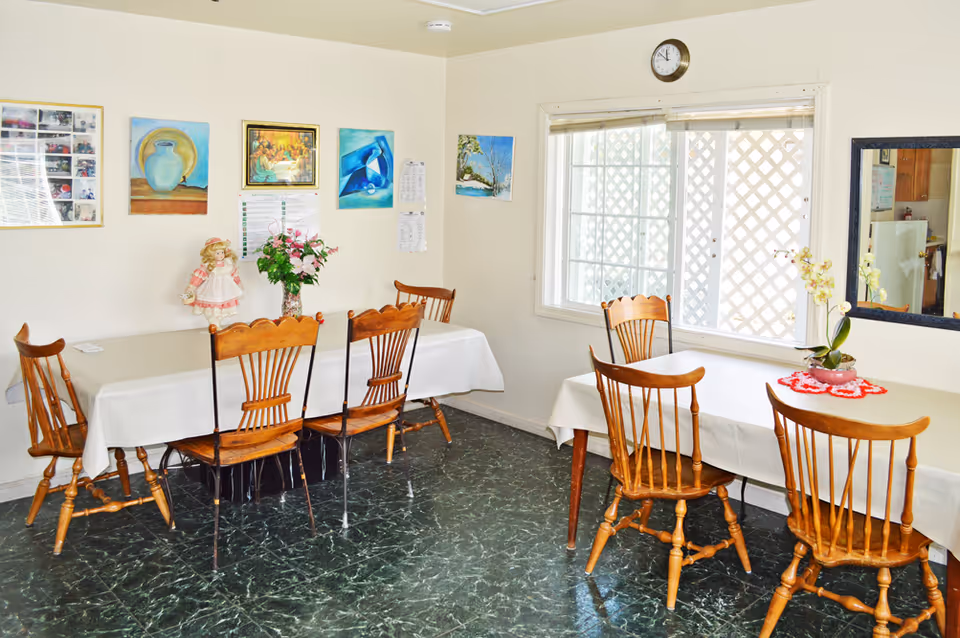 A dining room with two tables covered in white tablecloths, each surrounded by wooden chairs. The walls are decorated with framed pictures and paintings. A clock is mounted above a window with blinds, and a mirror hangs on the adjacent wall. There are flower arrangements on both tables.