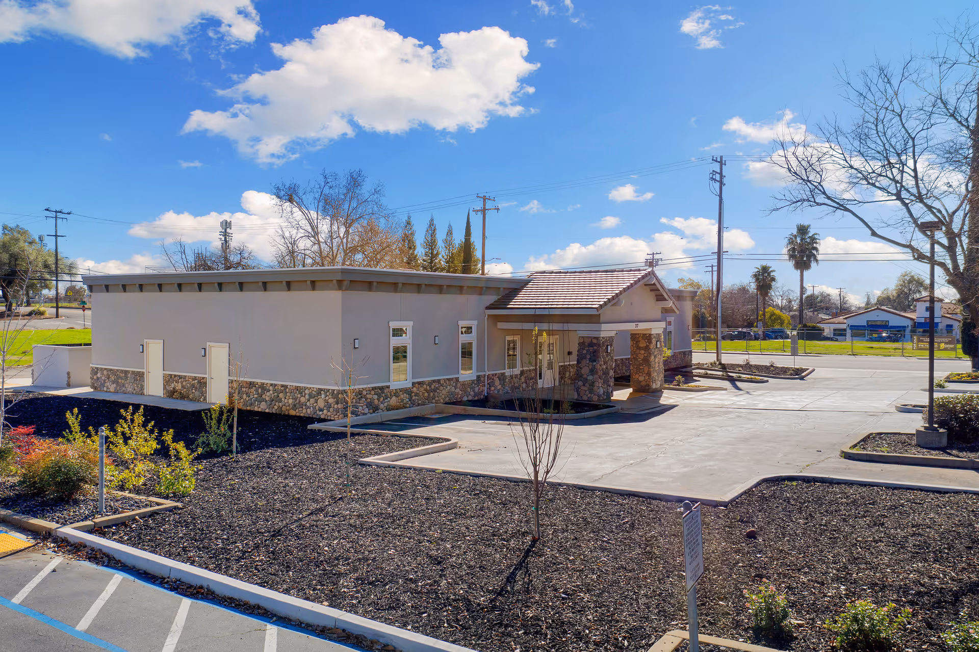 Exterior view of a single-story building with stone accents and a tiled roof entrance, surrounded by a parking lot and landscaped areas with small trees and shrubs under a partly cloudy blue sky.