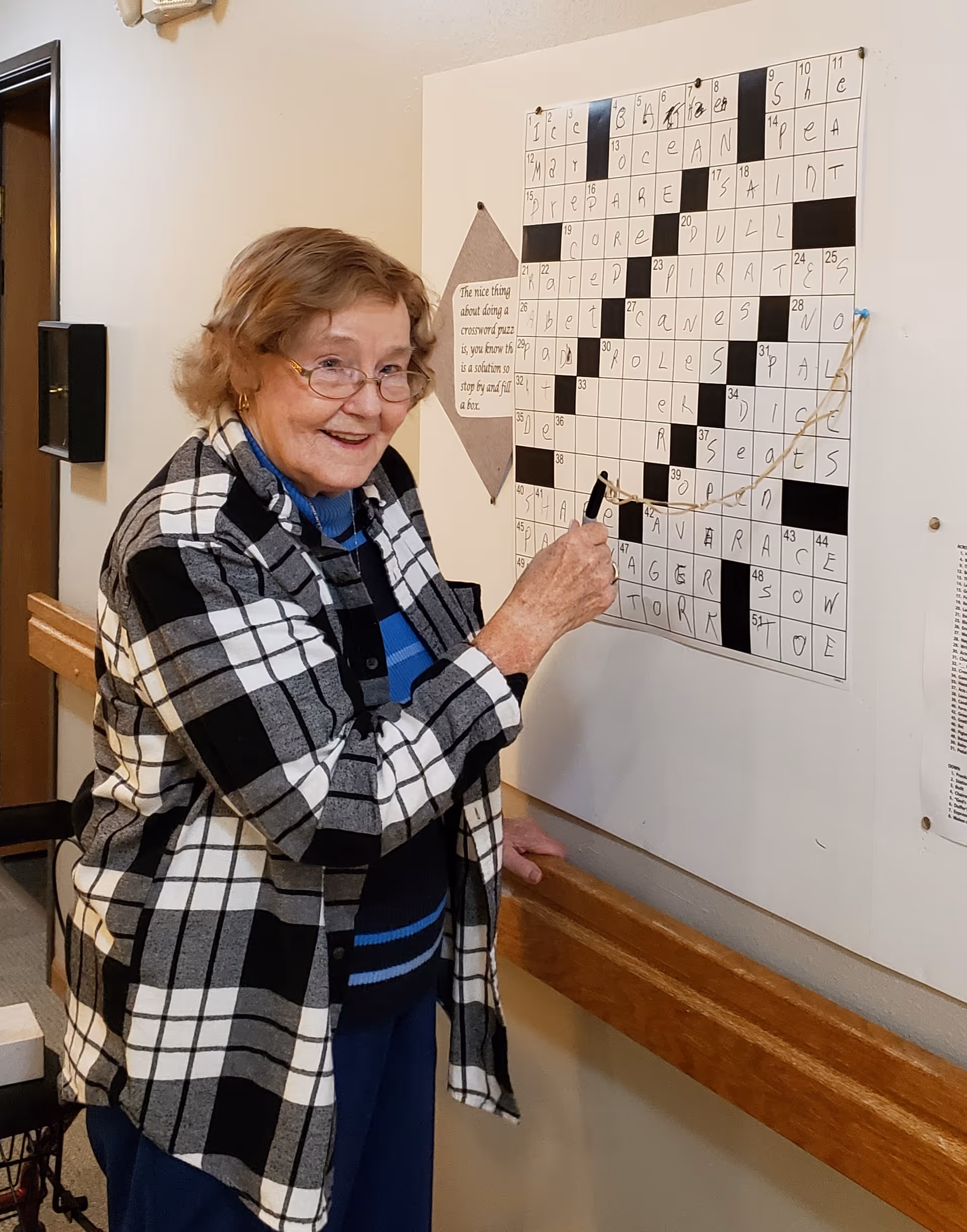 An elderly woman wearing glasses and a black and white checkered jacket is standing in a hallway, smiling and holding a marker while filling in answers on a large crossword puzzle mounted on the wall.
