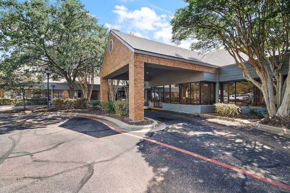Exterior view of Morada Temple senior living facility showing a covered entrance with brick pillars, large windows, surrounding trees, and a driveway with a red curb.
