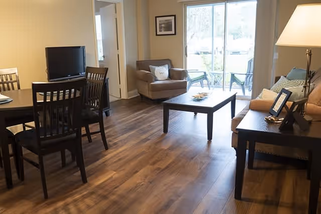 A cozy living room and dining area with wooden flooring. The living room has a beige sofa with cushions, a brown armchair, a coffee table, and a side table with a lamp and picture frame. The dining area features a dark wooden dining table with four chairs. Large sliding glass doors lead to an outdoor patio with chairs visible outside.