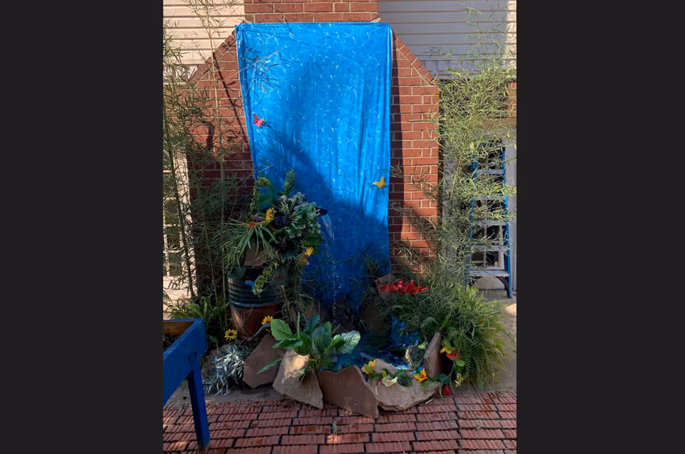 Outdoor garden display against a brick wall featuring a blue backdrop with butterfly decorations, surrounded by various green plants and flowers arranged with rocks on a brick-paved ground.