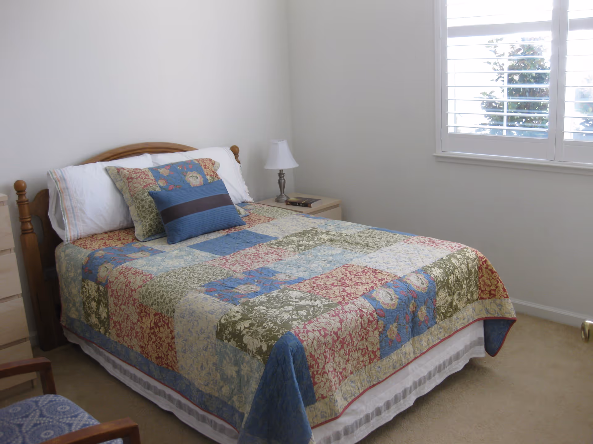A bedroom with a wooden bed frame and a colorful patchwork quilt on the bed. There are three pillows on the bed, including a blue decorative pillow. A small nightstand with a lamp and a book is next to the bed. A window with white shutters lets in natural light, and a wooden chair with a blue patterned cushion is partially visible.