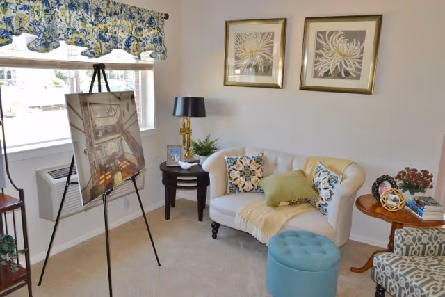 A cozy living room area with a white tufted loveseat adorned with floral and green pillows and a yellow throw blanket. A blue round ottoman is placed in front of the loveseat. To the right, there is a wooden side table with a decorative globe, books, and a vase of flowers. On the left side, a small round table holds a lamp and a small plant. A large window with floral curtains lets in natural light, and an easel displaying a framed picture stands near the window. Two framed floral artworks hang on the wall above the loveseat.