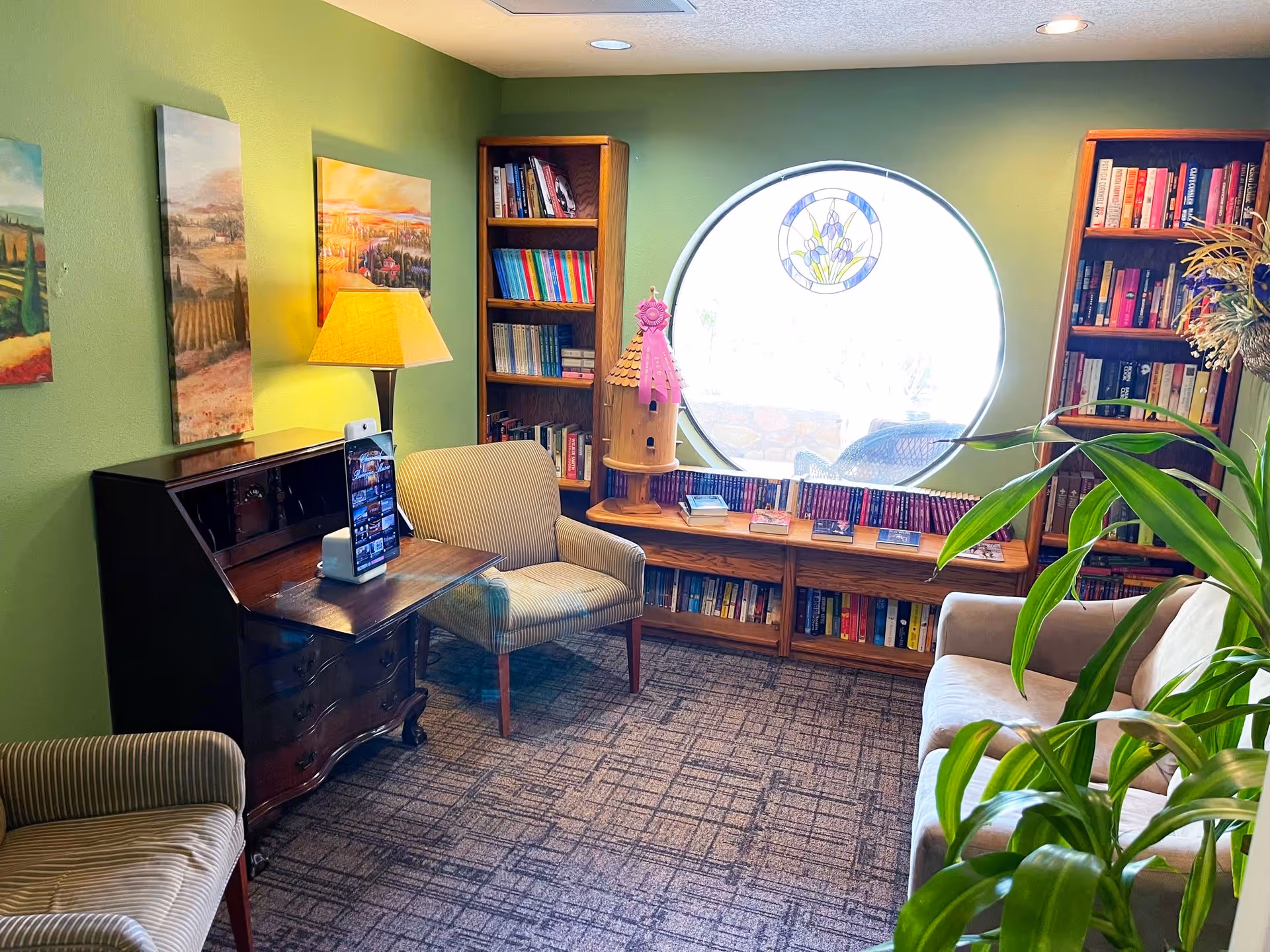 A cozy living room area with green walls, two bookshelves filled with books, a round window with a stained glass decoration, a wooden desk with a lamp and a tablet on a stand, two striped armchairs, a beige sofa, and a large green plant in the foreground.