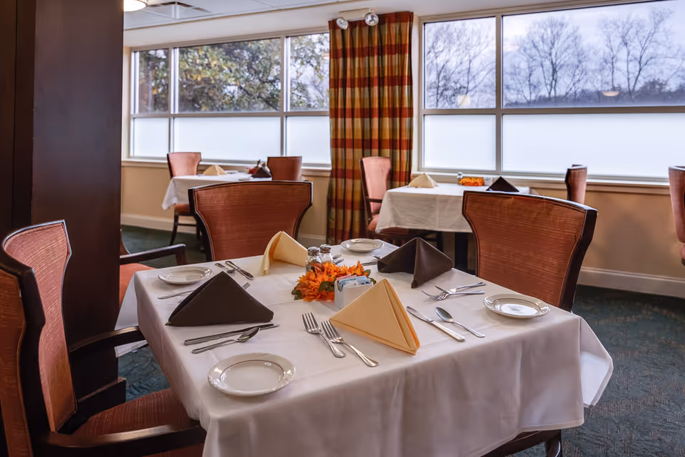 A dining room with tables set for a meal, featuring white tablecloths, folded napkins in beige and brown, plates, silverware, and a small floral centerpiece. The room has large windows with frosted lower panes and red and yellow plaid curtains, and several chairs with reddish upholstery around the tables.