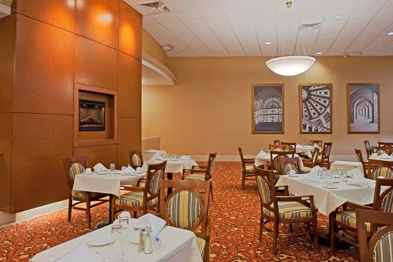 Dining room with multiple tables covered in white tablecloths, set with glassware, napkins, and silverware. The room features a patterned red carpet, wooden chairs with striped cushions, a wall-mounted fireplace, and three framed architectural photographs on the beige walls. A modern ceiling light fixture hangs above.