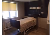 A small, simple bedroom in a rehabilitation and nursing facility featuring a single hospital-style bed with a beige quilted blanket. The room has a window with a closed beige roller shade, a wall-mounted reading light above the bed, and a wooden floor. The walls are painted in neutral tones with a dark accent wall behind the bed.