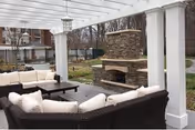 Outdoor covered patio with wicker sectional seating and a stone fireplace beneath a white pergola.