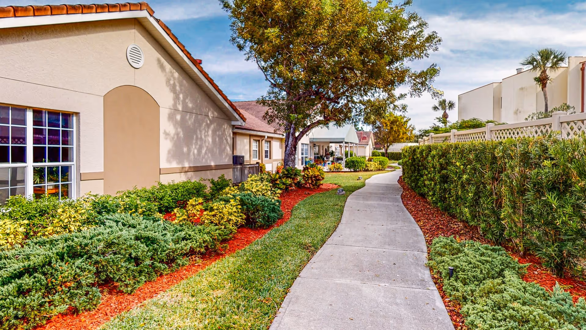A paved walkway curves through a landscaped garden area with green shrubs, bushes, and trees on both sides. On the left side, there is a beige building with a red-tiled roof and windows. The right side features a tall hedge and a white fence. The sky is partly cloudy with blue patches.