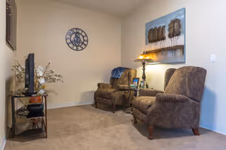 A cozy interior living area with two patterned armchairs, a small side table with a lamp and framed photo, a wall clock, a decorative painting of trees, and a TV on a stand with shelves holding decorative items.