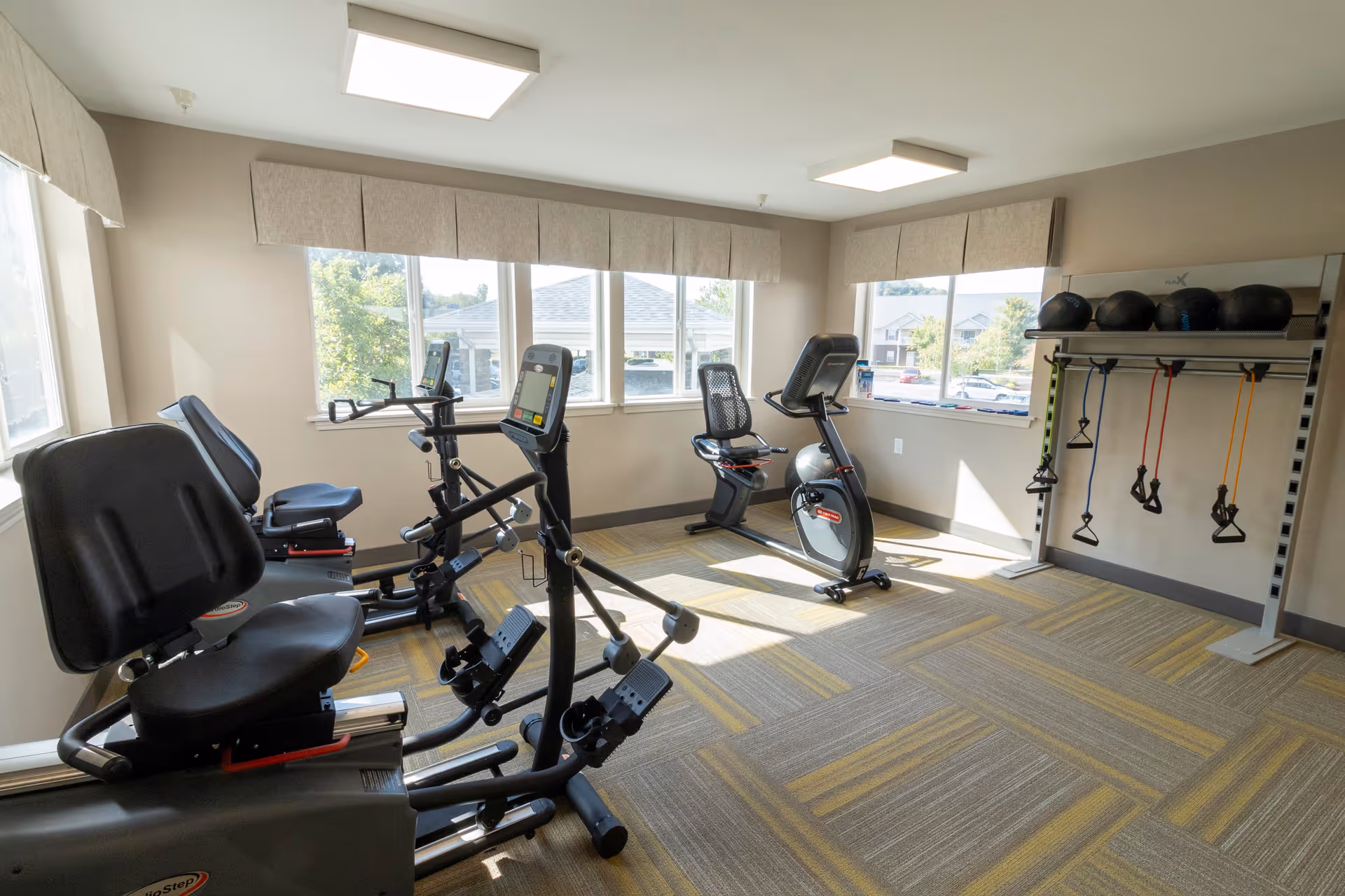 A bright exercise room with large windows showing an outdoor view. The room contains several stationary exercise bikes and a rack with resistance bands and medicine balls. The floor is carpeted with a patterned design, and the walls are painted light beige.