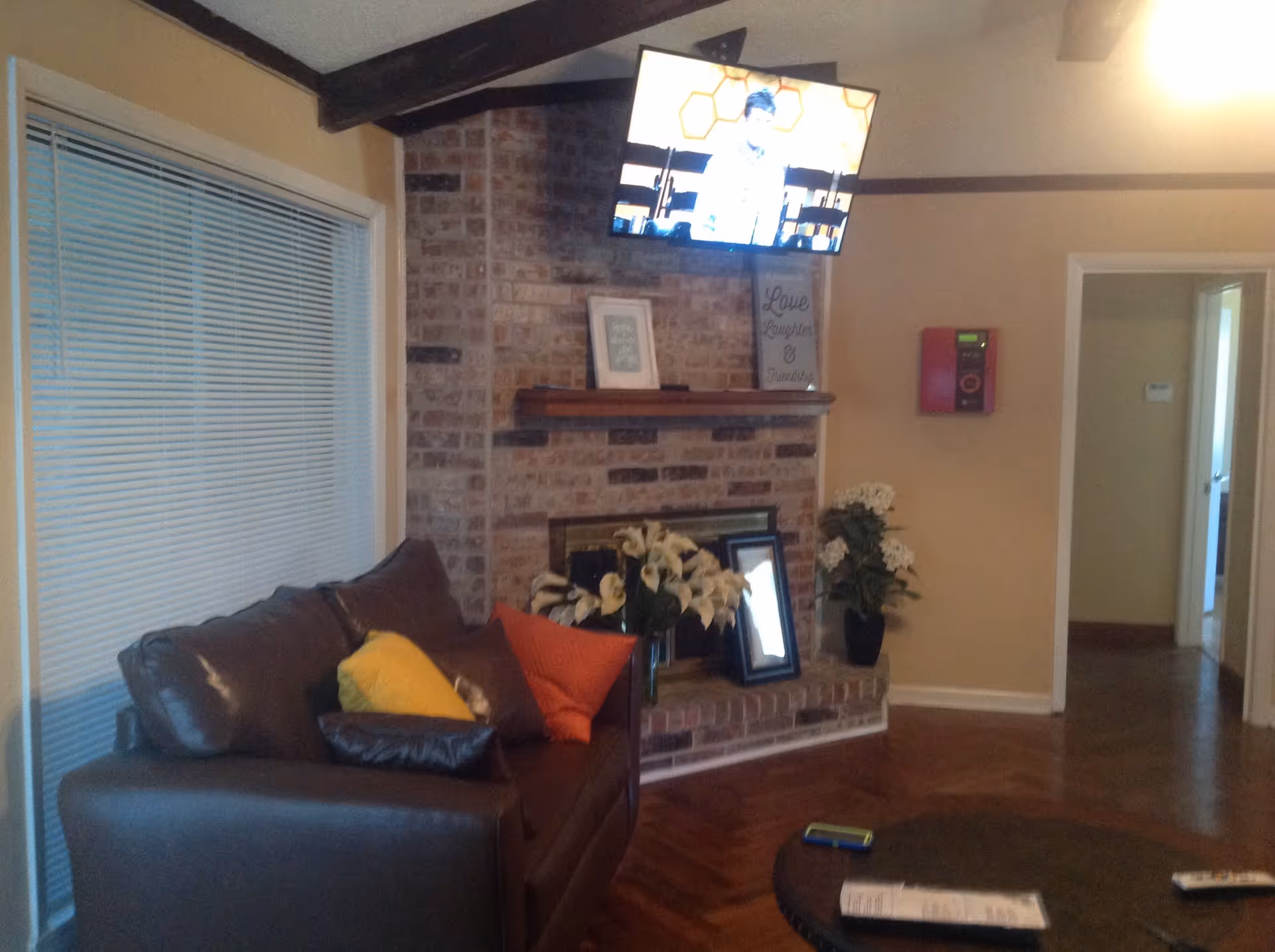 A cozy living room with a brown leather couch adorned with colorful pillows, a brick fireplace decorated with framed pictures and flower vases, a wall-mounted TV above the fireplace, and a round coffee table with a remote and papers on it. There is a window with blinds on the left and a doorway leading to another room on the right.