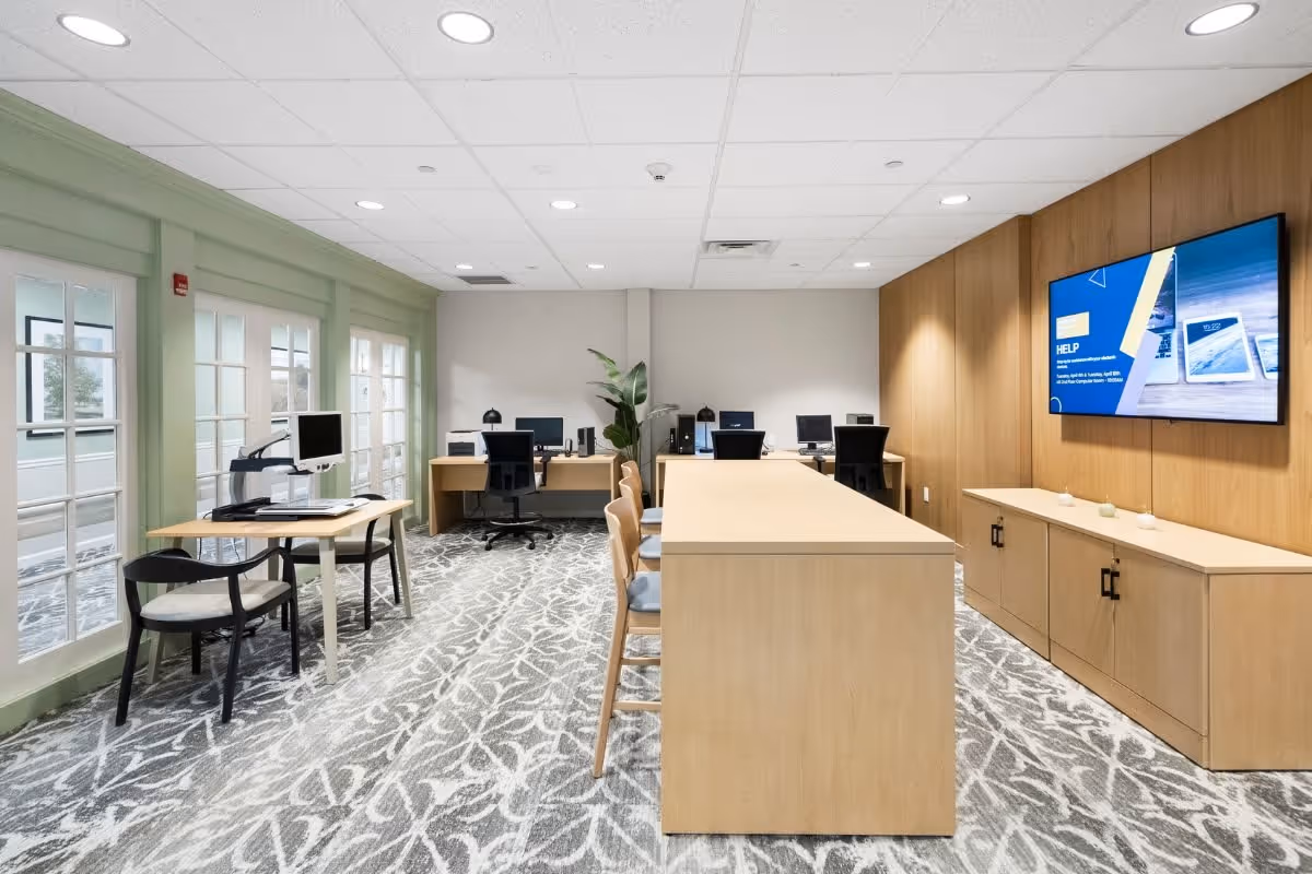 A modern office space with a long wooden table in the center surrounded by chairs. Along the back wall, there are desks with computer monitors and office chairs. The right wall features wood paneling with a large flat-screen TV mounted on it. The left side has large windows with green trim and a small table with two chairs. The floor is carpeted with a gray and white patterned design.