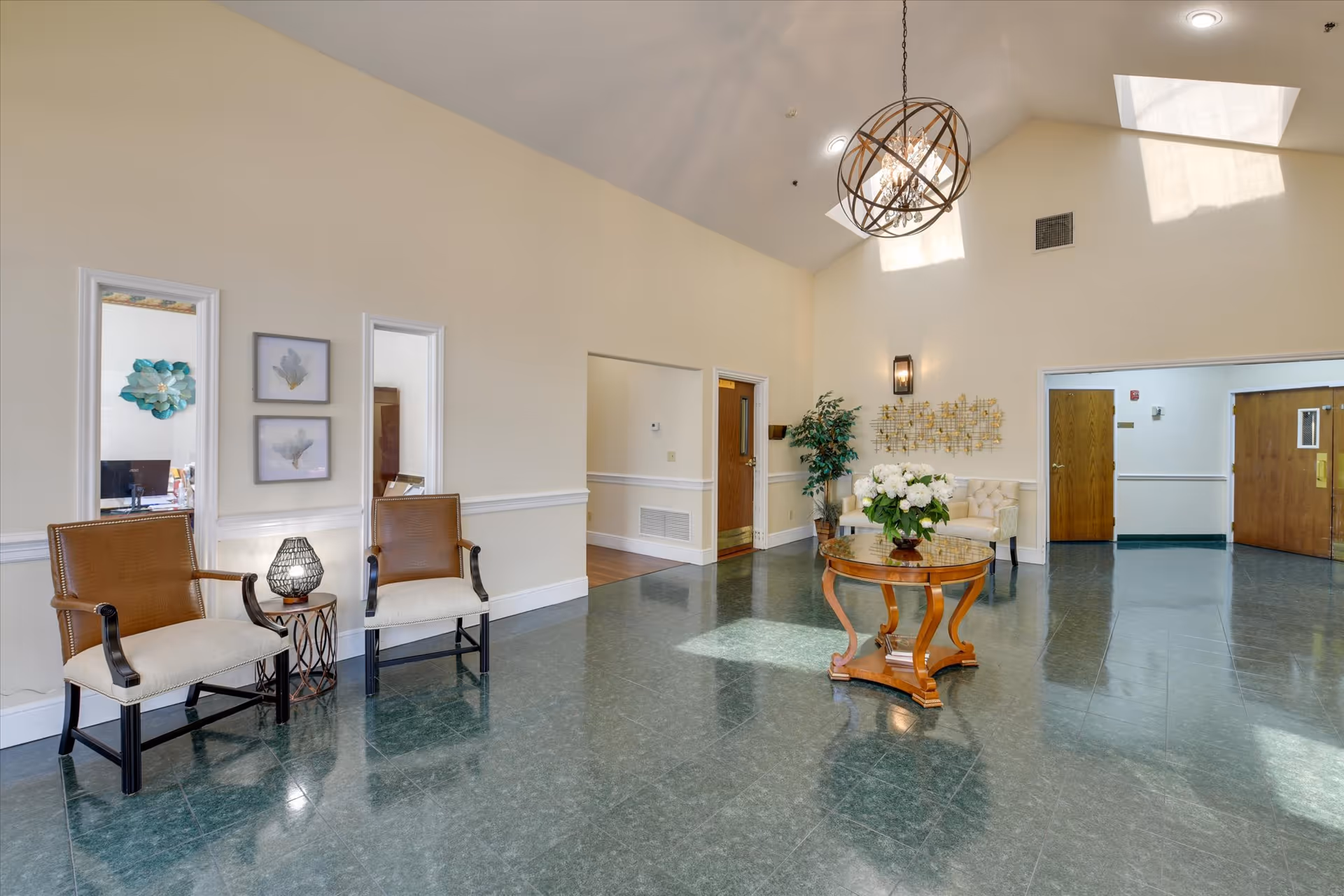 A spacious and well-lit interior common area with high ceilings and skylights. The room features a round wooden table with a floral centerpiece, two brown and beige armchairs with a small side table and lamp between them, a cream-colored tufted bench, wall art, and a potted plant. There are multiple doorways leading to other rooms, and the floor is covered with greenish tiles.
