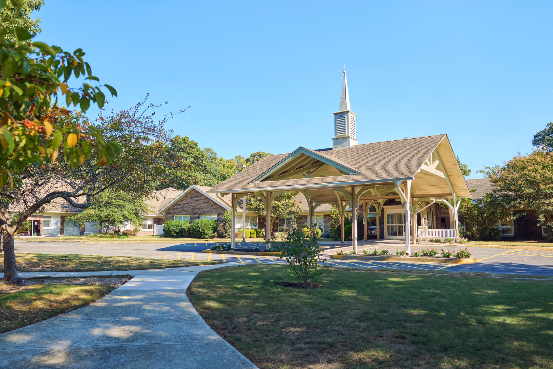 Exterior view of Pelican Pointe Post Acute Nursing & Rehabilitation facility featuring a covered entrance with a peaked roof and a small steeple. The building is surrounded by trees and greenery under a clear blue sky, with a paved walkway leading to the entrance.
