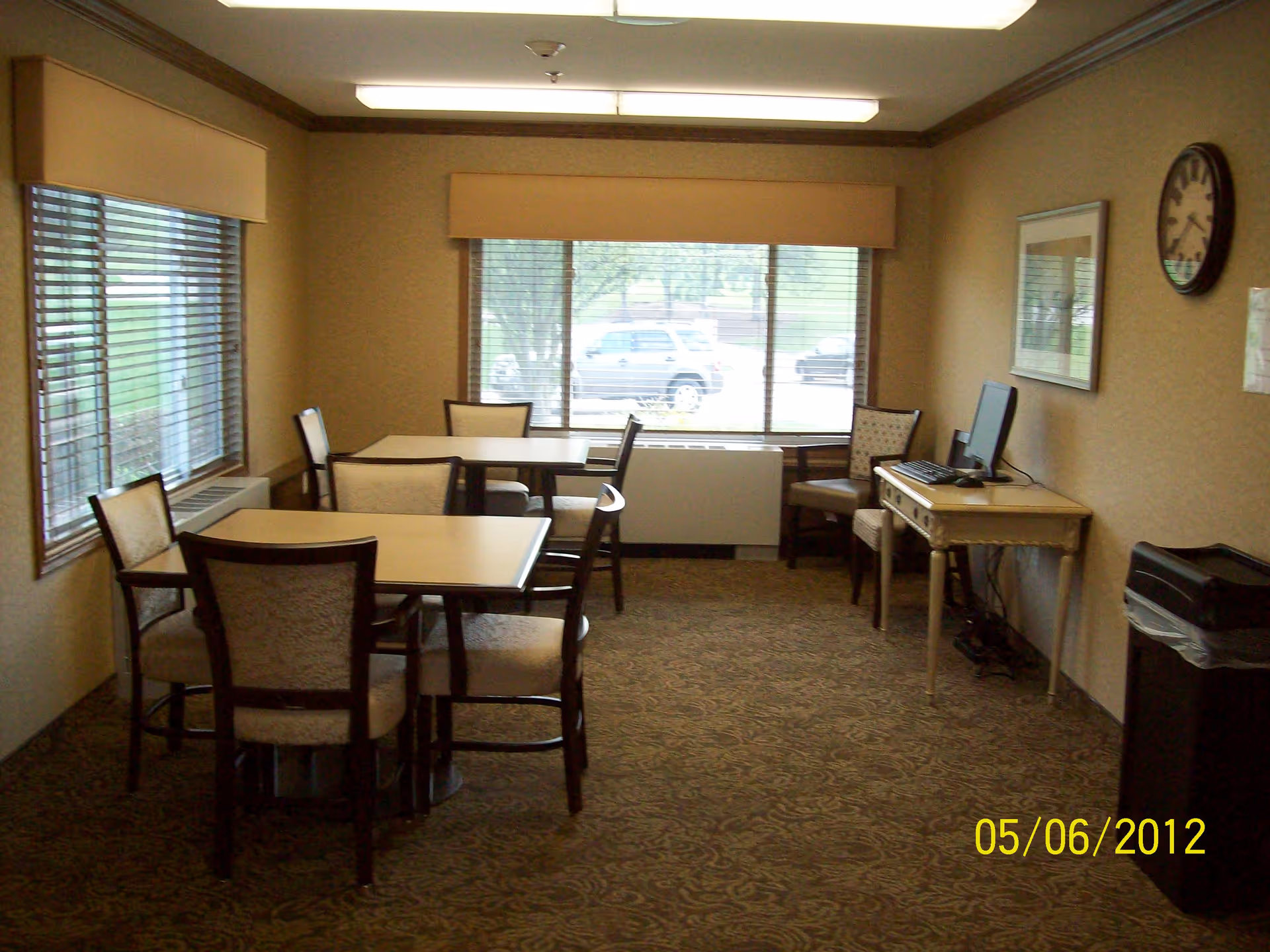 A small room with two tables and several chairs arranged around them. There is a large window with blinds letting in natural light. On the right side, there is a small desk with a computer and a chair, a wall clock above it, and a trash can nearby. The room has patterned carpet and beige walls.