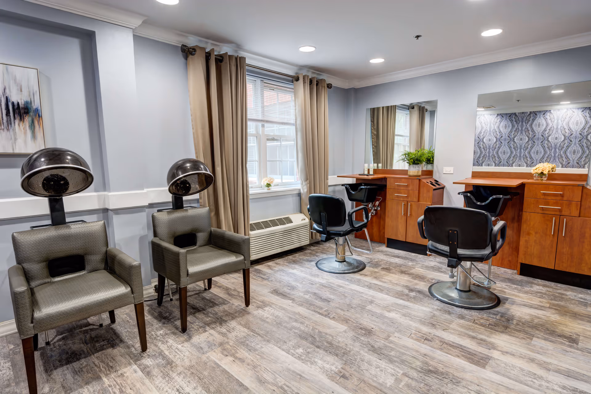 Interior view of a salon area with two hair drying chairs on the left and two styling chairs in front of wooden cabinets with mirrors. The room has light blue walls, beige curtains on the window, and wood-patterned flooring.