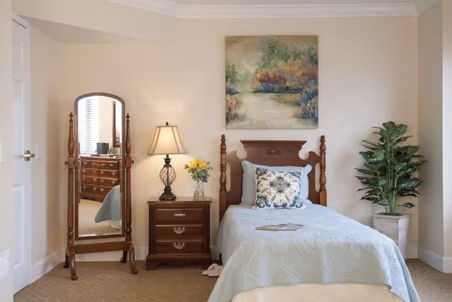 A cozy bedroom featuring a single wooden bed with a light blue quilt and decorative pillows. Next to the bed is a wooden nightstand with a lamp and a vase of yellow flowers. A full-length wooden framed mirror stands to the left of the nightstand, reflecting part of a dresser. A potted plant is placed on the right side of the bed, and a landscape painting hangs on the wall above the headboard. The room has beige walls and carpeted flooring.