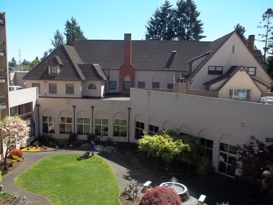 Sunlit courtyard with a green lawn, walking paths, a small fountain, and a surrounding multi-story retirement building.