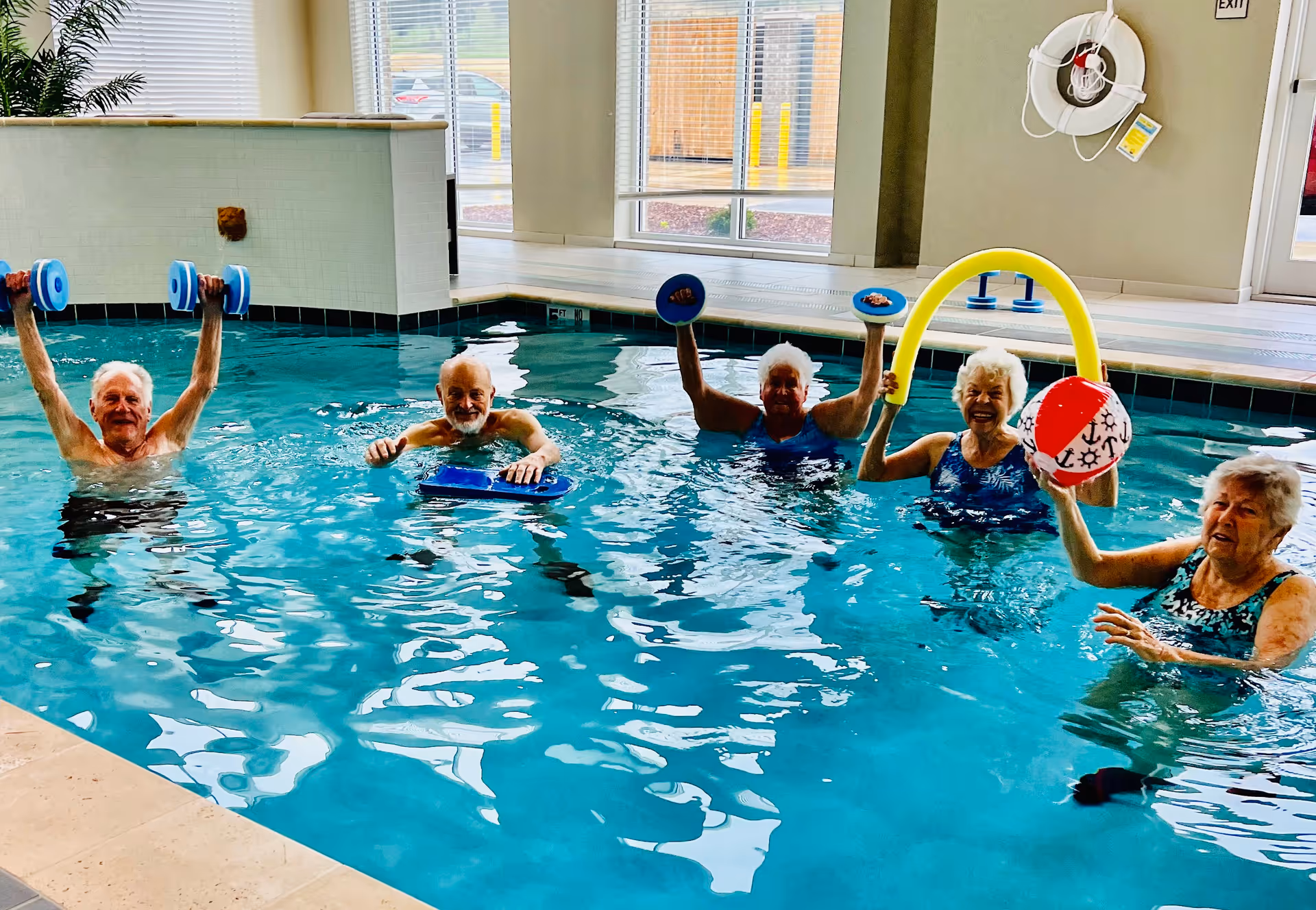 Five elderly individuals participating in a water exercise class in an indoor swimming pool. Two are holding blue dumbbells above the water, one is using a blue kickboard, one is holding a yellow pool noodle and a red and white beach ball, and another is reaching out with her hand. The pool area has large windows letting in natural light and a life preserver mounted on the wall.