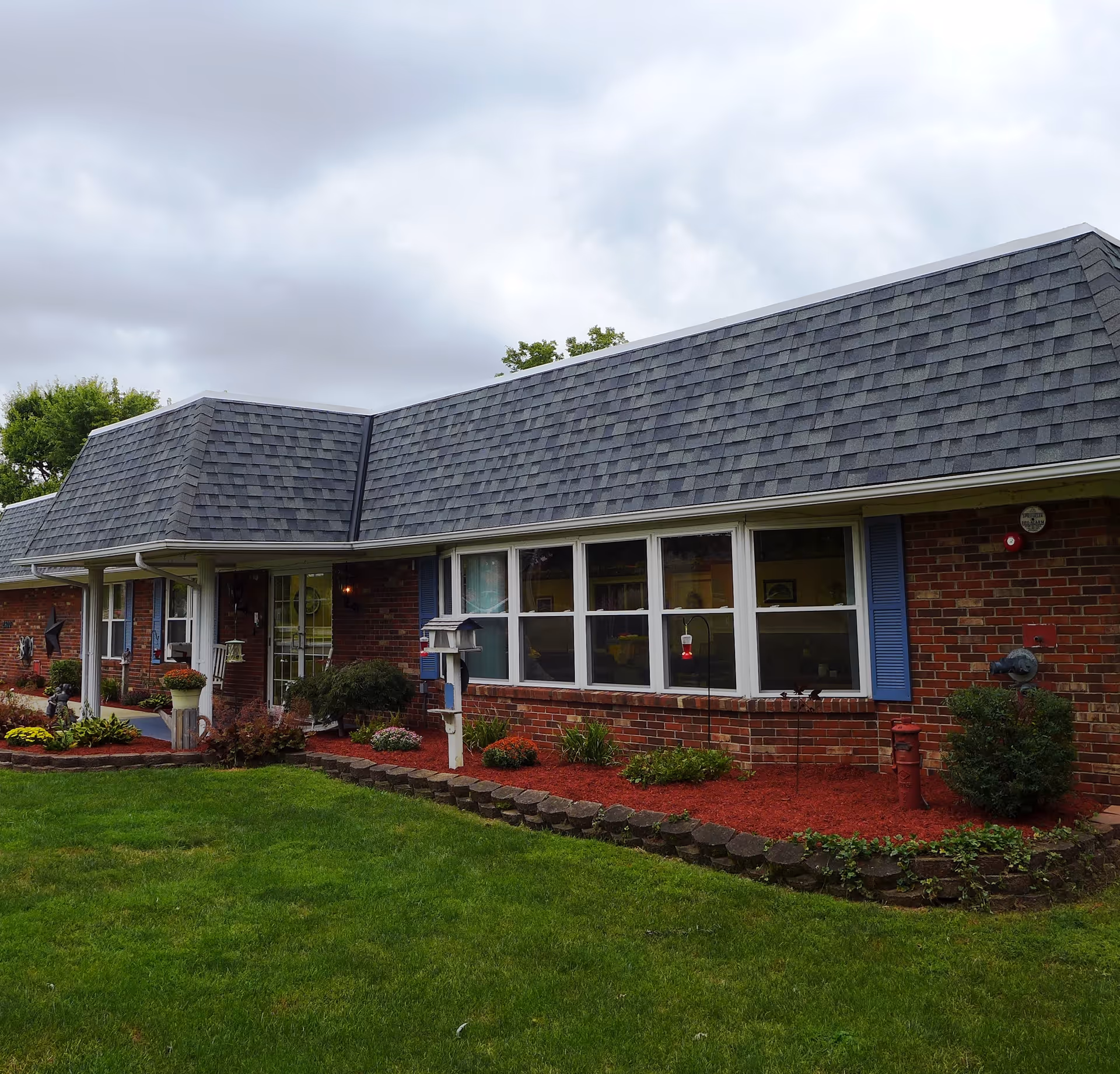 Exterior view of a single-story brick building with a gray shingled roof, blue shutters, and several windows. The building is surrounded by a well-maintained lawn and landscaped garden beds with red mulch, shrubs, and flowers. The sky is overcast.