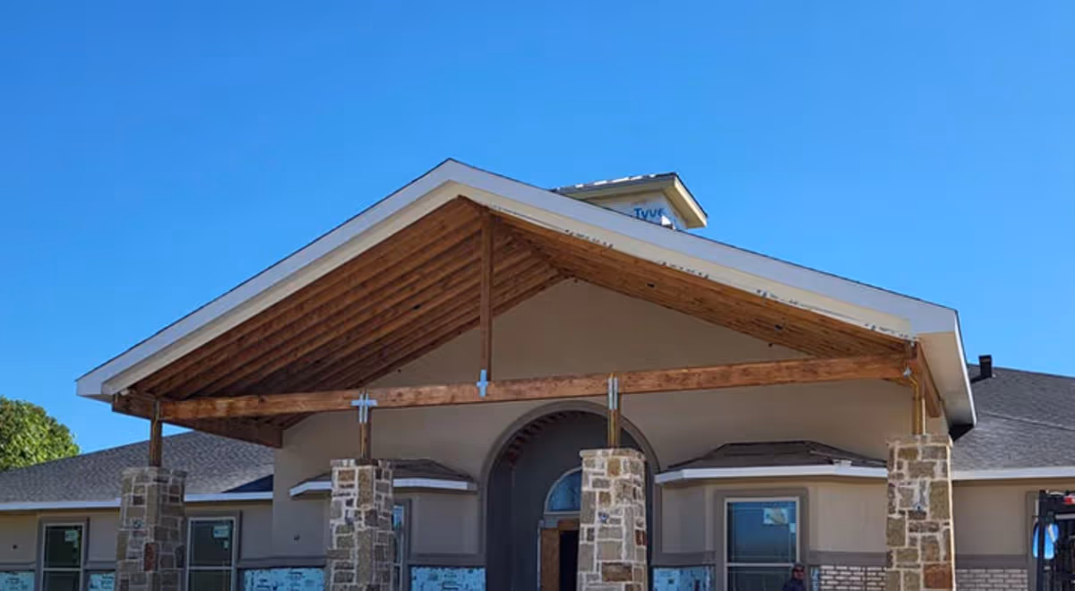 Front exterior view of a building under construction with a large wooden gabled roof supported by stone pillars, clear blue sky in the background.