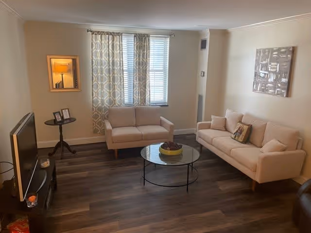 A cozy living room with two beige sofas facing each other, a round glass coffee table with a decorative bowl in the center, a TV on a stand to the left, patterned curtains on the window, a small side table with framed photos, and a wall mirror reflecting a lamp.