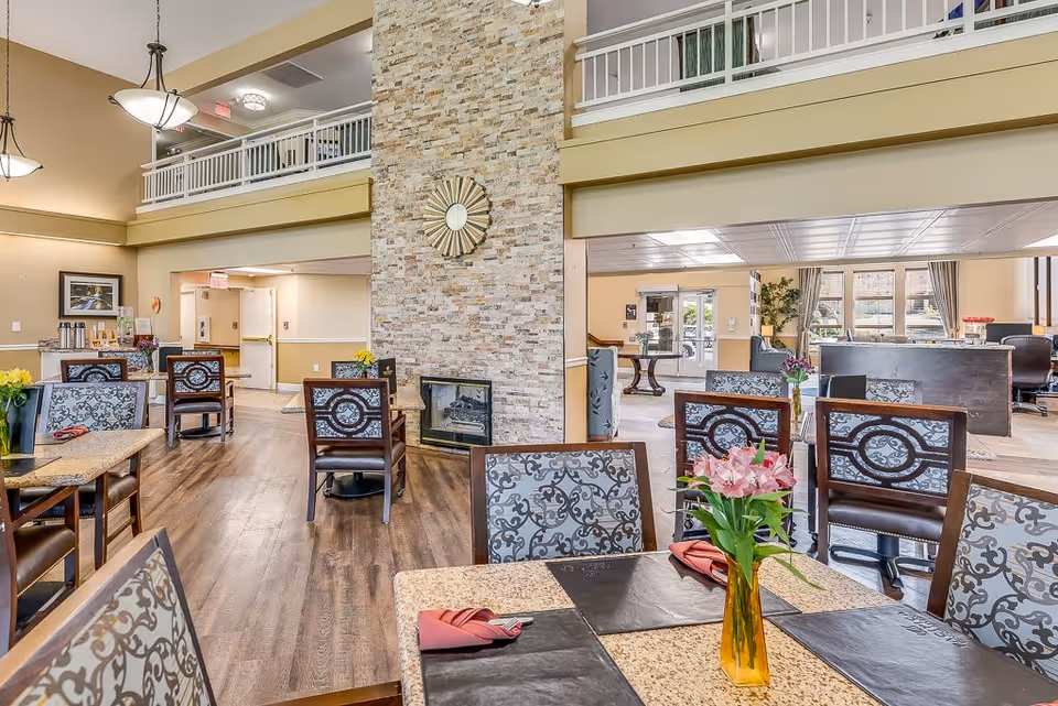 A spacious and well-lit dining area in a senior living facility with multiple tables and chairs. The tables are set with placemats, napkins, and a vase with flowers. There is a stone fireplace in the center with a decorative mirror above it. The room has high ceilings with hanging light fixtures and an upper balcony railing. In the background, there is a reception or common area with large windows and additional seating.