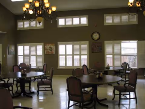 Senior living facility dining room with round tables, chairs, chandeliers, and large windows.