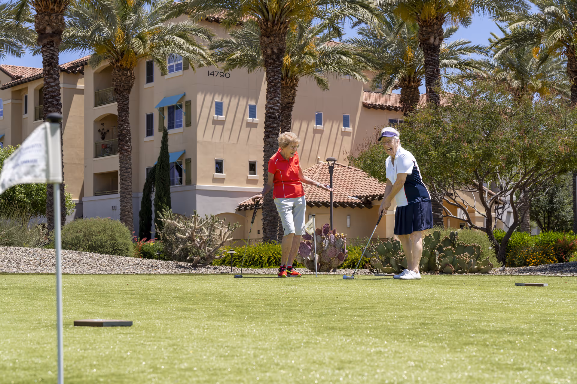 Two older adults putting on a green near palm trees with a Mediterranean-style apartment building in the background.