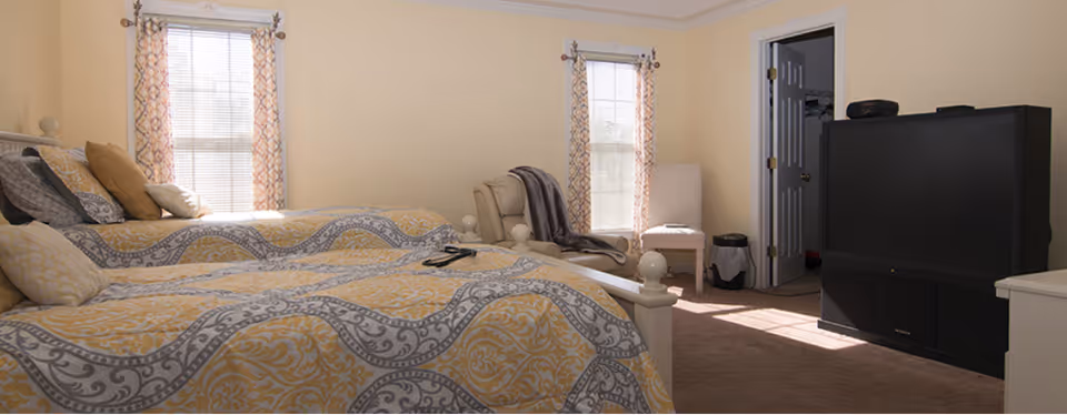 A bright bedroom with two twin beds dressed in yellow-and-gray patterned bedding, two windows, an armchair, and a large television.