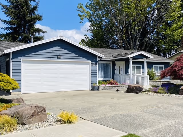 Single-story residential building with blue siding and a white garage door, surrounded by trees and landscaped with rocks, bushes, and flowers under a partly cloudy sky.