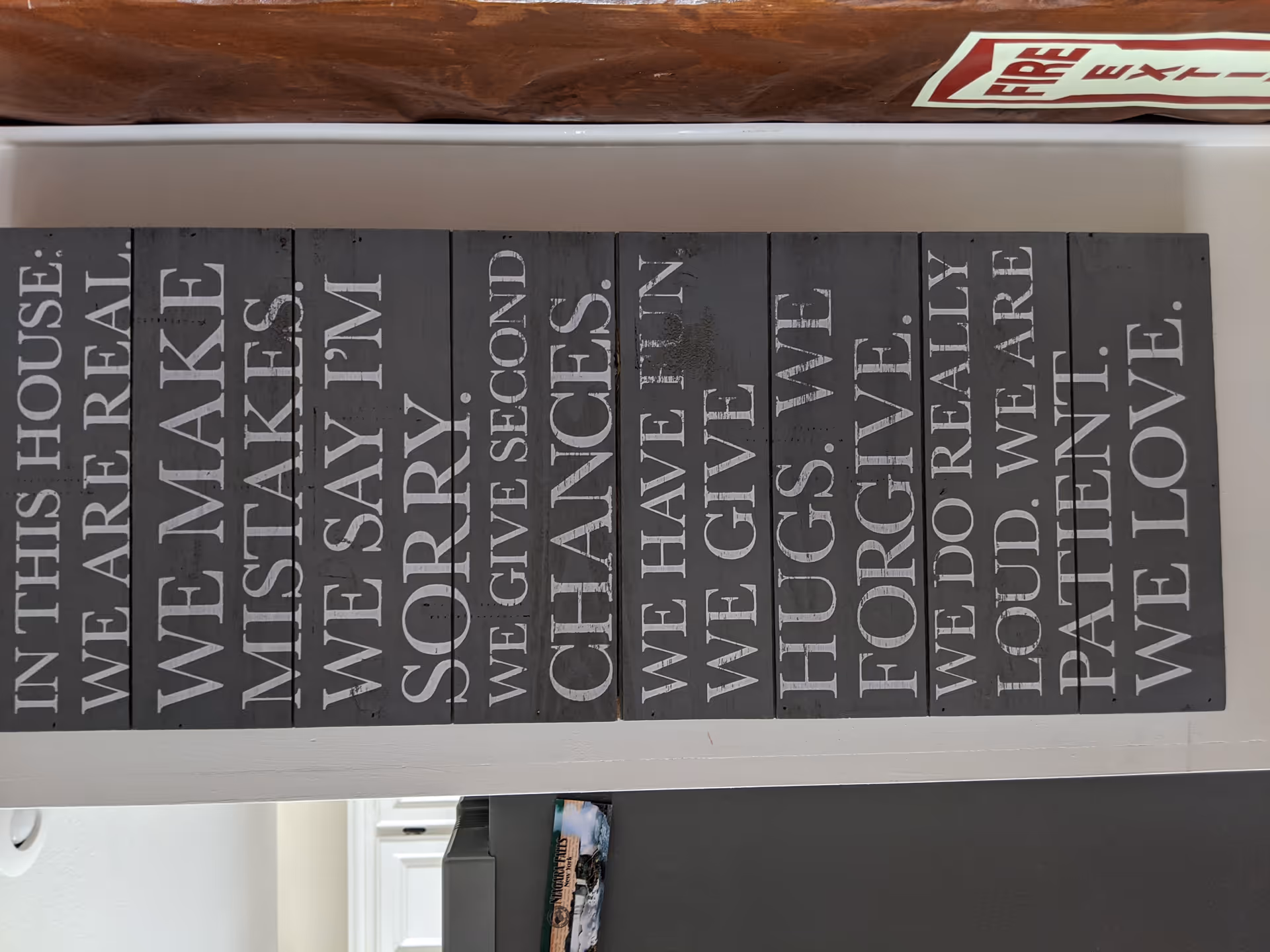 A decorative wall sign with multiple wooden panels painted black and white text listing family values and rules such as 'In this house we are real', 'We make mistakes', 'We say I'm sorry', 'We give second chances', 'We have fun', 'We give hugs', 'We forgive', 'We do really loud', 'We are patient', and 'We love'. The sign is mounted on a white wall with a wooden beam above it and a fire extinguisher sign partially visible at the top.