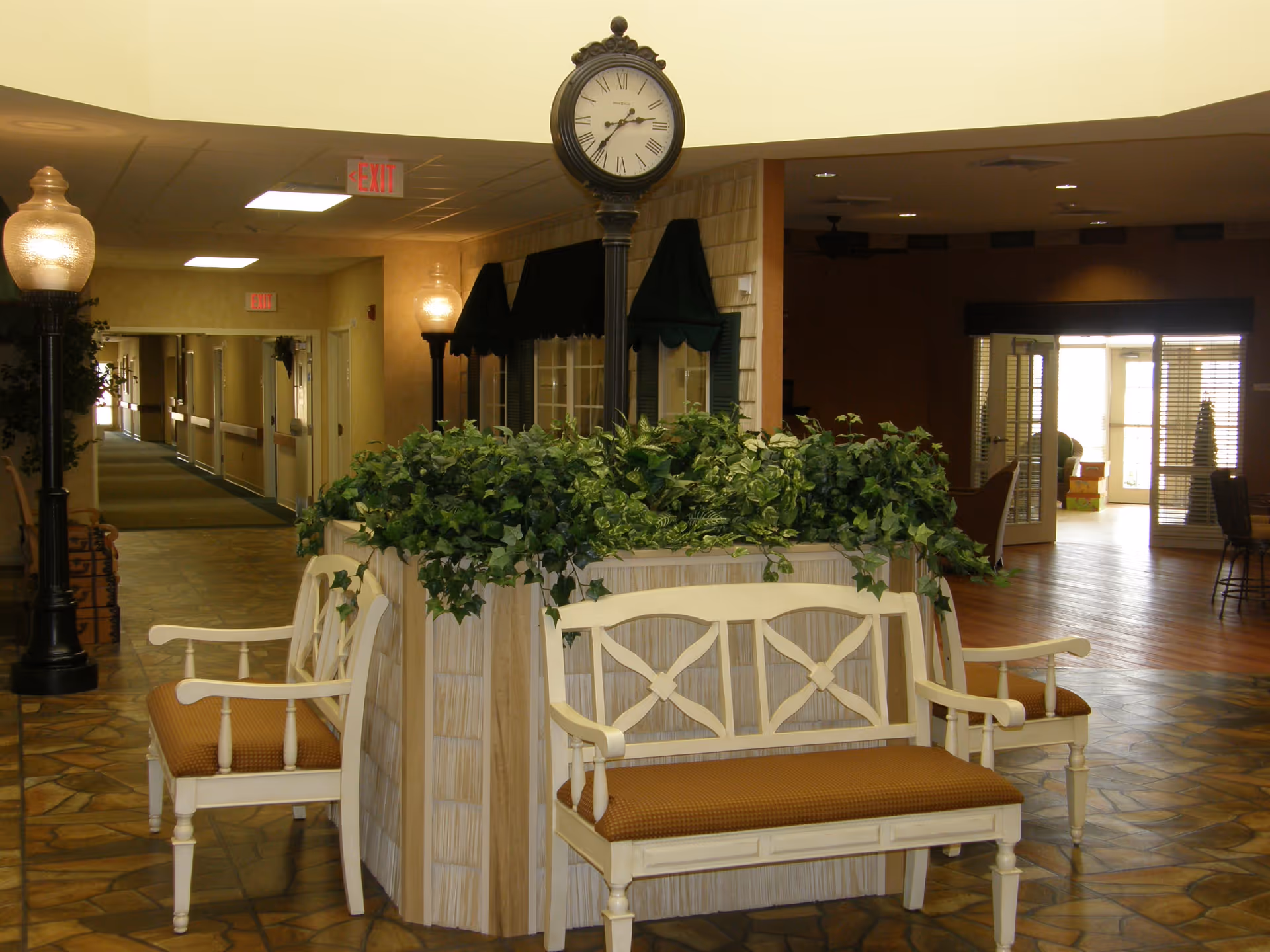 Indoor seating area with cream-colored wooden benches with brown cushions surrounding a planter filled with green leafy plants. A tall black clock with Roman numerals stands in the center of the planter. The area has tiled flooring, warm lighting, and a hallway leading to other rooms. There are also two vintage-style lamp posts on either side of the planter.