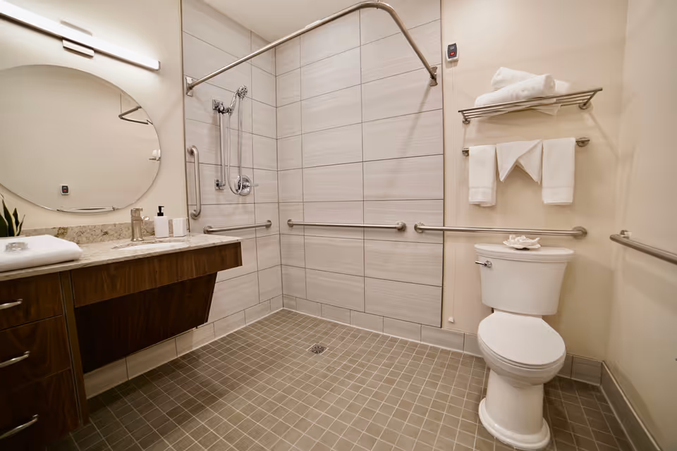 A modern, accessible bathroom with a walk-in shower area featuring grab bars and a handheld showerhead. The bathroom includes a white toilet with a towel rack above it holding folded white towels. There is a wooden vanity with a granite countertop, a soap dispenser, and a large oval mirror above it. The floor is tiled with small square tiles, and the walls have larger rectangular tiles in the shower area.